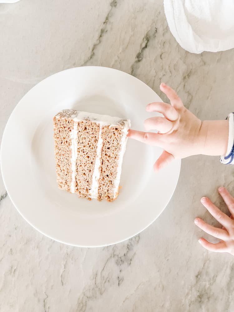 a slice of carrot cake on a white plate with toddler hands touching the frosting
