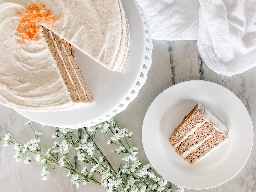 a slice of carrot cake with marshmallow cream cheese frosting on a white plate with a tea towell and flowers